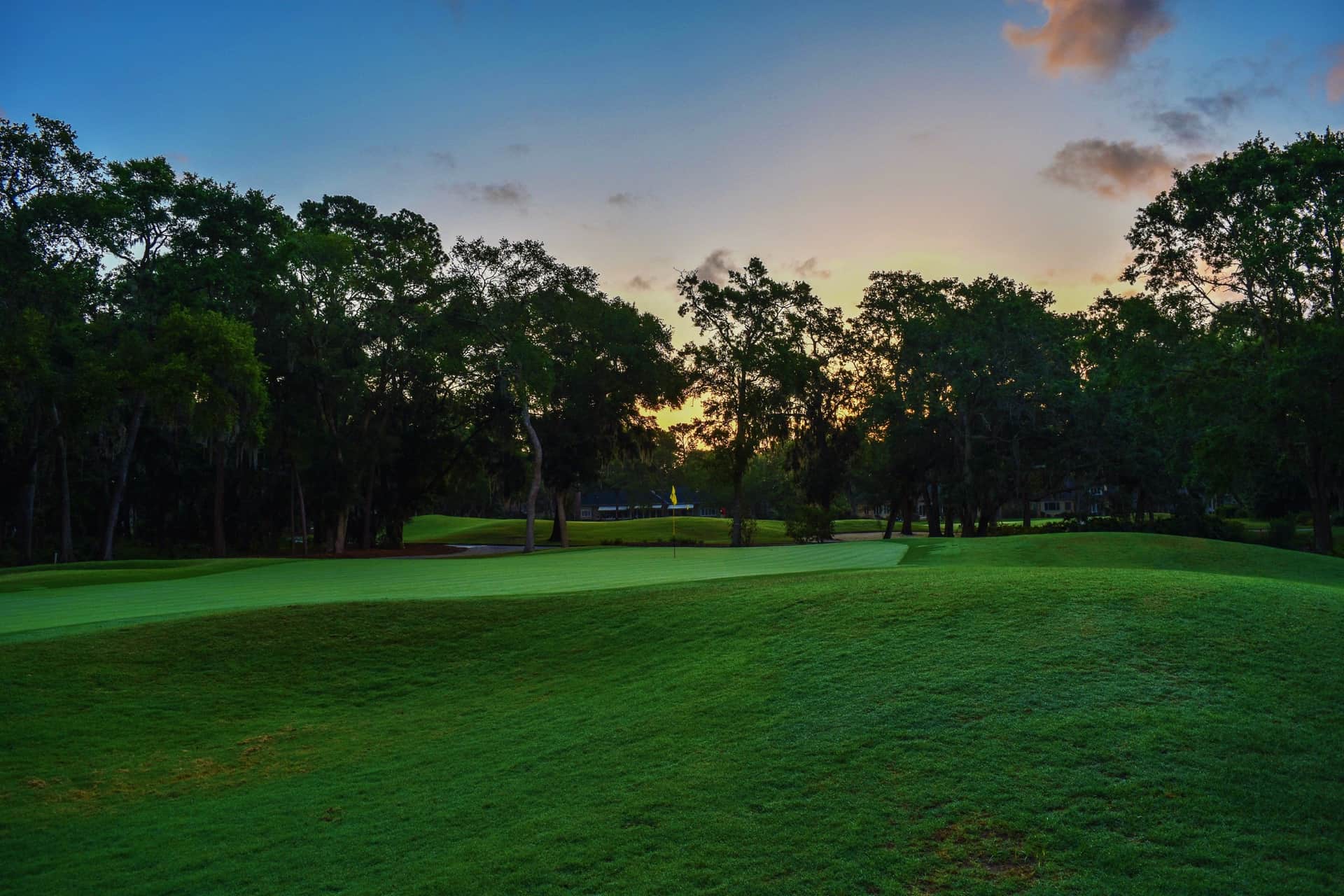 A manicured golf green with the flagstick at dusk, the clubhouse beyond the trees